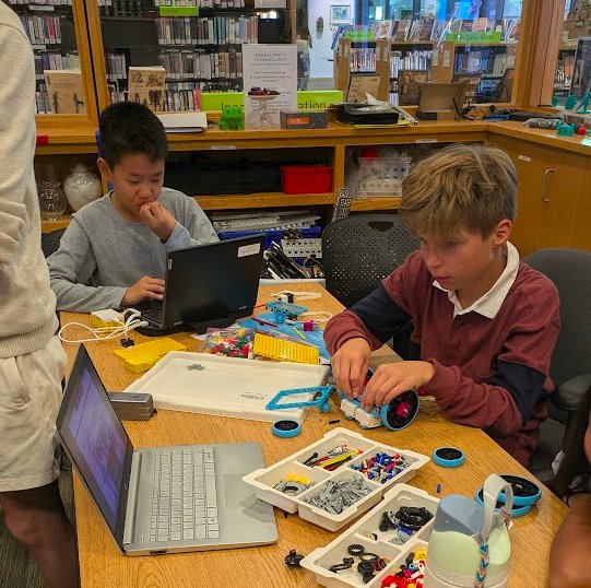 Two students building a LEGO robot at a library table, laptops open